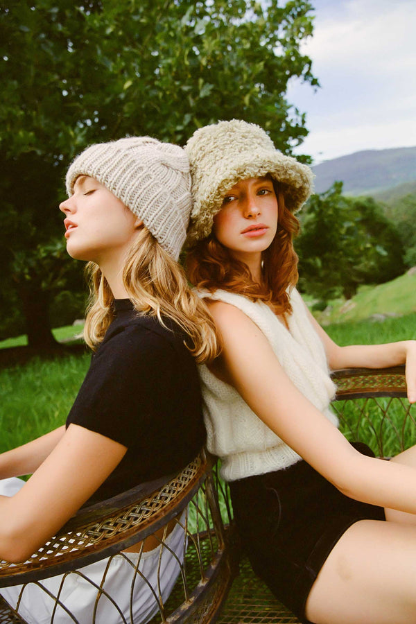 Two models wearing bucket hats, one in fluffy sage green and the other in beige, sitting outdoors.