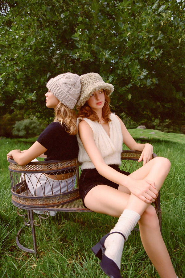 Two models wearing fluffy bucket hats, one in sage green, sitting on a bench in a grassy area.