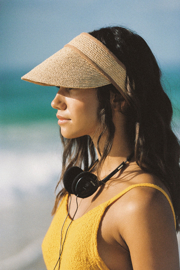 Profile view of a woman wearing a natural straw linen visor at the beach, with headphones around her neck.