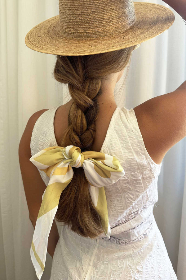 A woman with a braided hairstyle wearing a yellow and white scarf tied at the back of her head, complemented by a straw hat.