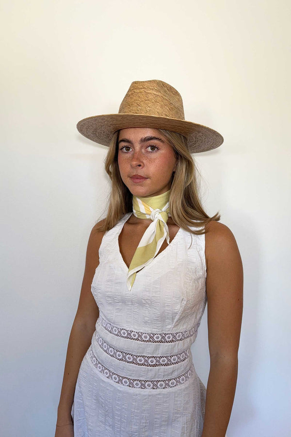 Model wearing a pistachio scarf tied around the neck, with a straw hat, in a white dress.