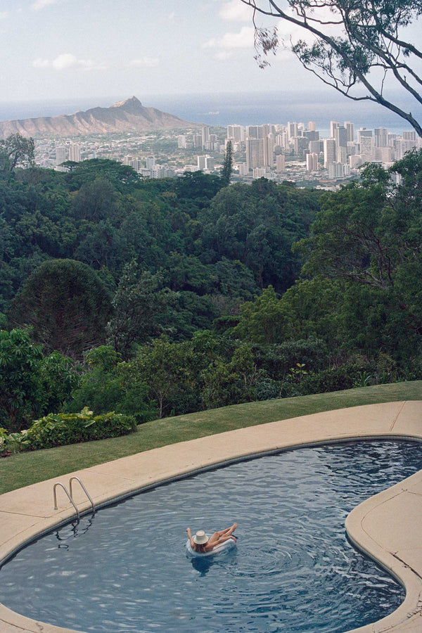 A person relaxing in a pool wearing a straw bucket hat, with a city skyline in the background.