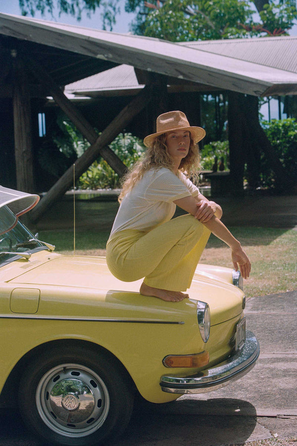 Model wearing The Inca Fedora, a natural straw hat, sitting on a yellow car