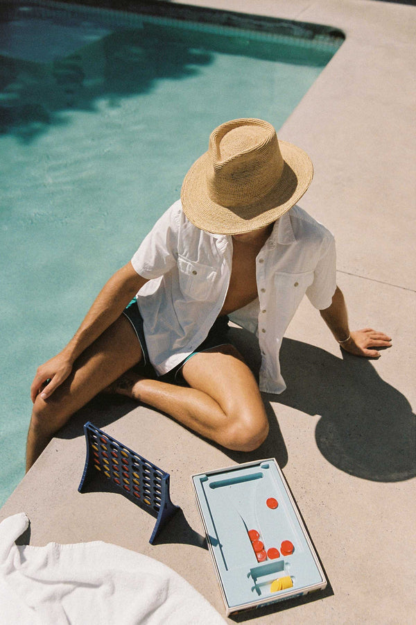 A person sitting by a pool wearing a natural straw fedora hat, with a game nearby.