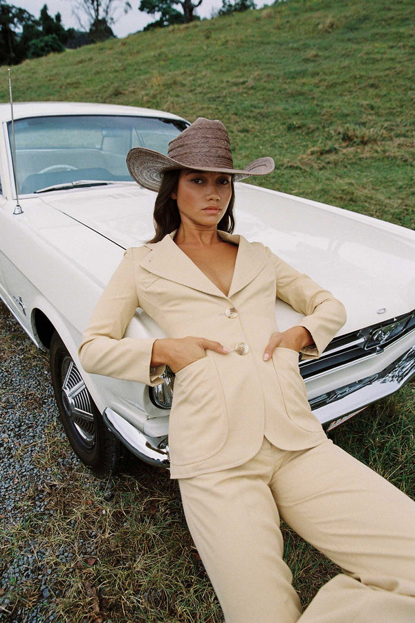 Model wearing a Wesley Cowboy straw hat in brown, styled with a beige suit, sitting on a white car.