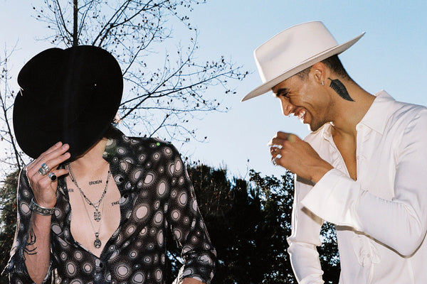 Two people outdoors, one wearing a white fedora hat, the other in a black hat, smiling and interacting.