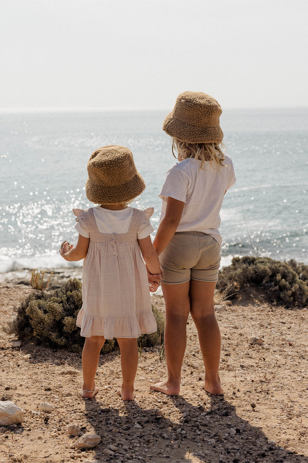 Two children holding hands at the beach, wearing brown Teddy Bucket hats.