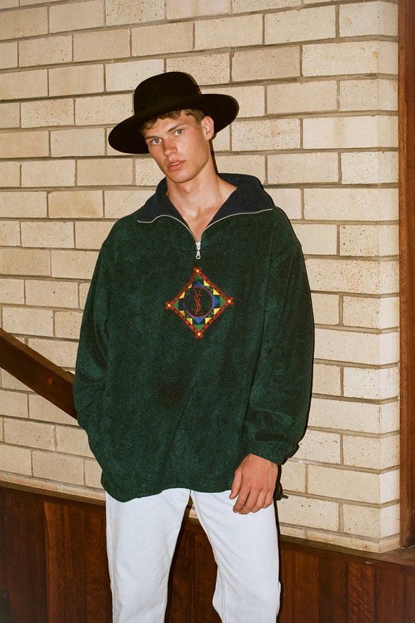 A model wearing a black wool felt fedora hat, styled with a green pullover and standing against a brick wall.