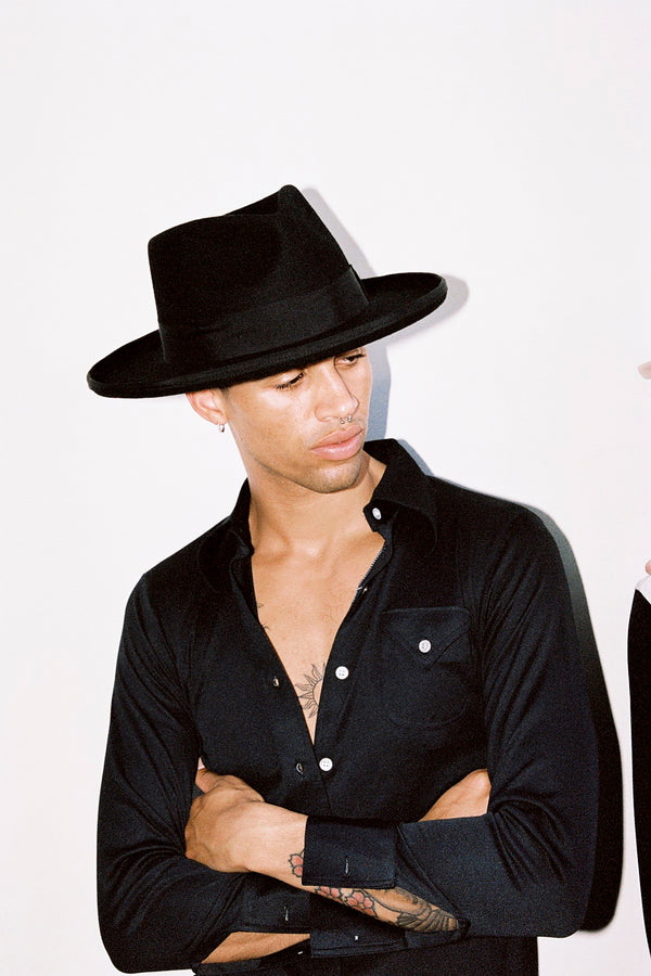 Model wearing a black wool felt fedora hat, arms crossed, with a neutral background.