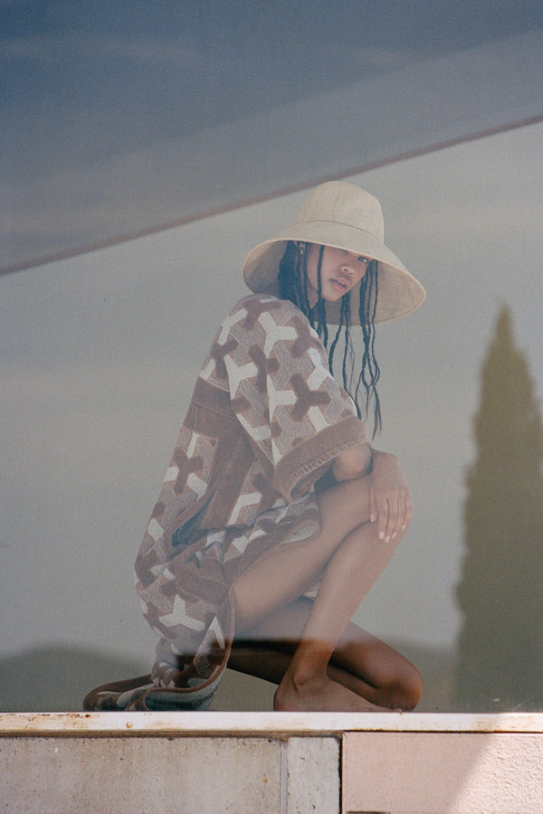Model wearing The Cove straw bucket hat, crouching by a reflective surface, wearing a patterned cover-up.
