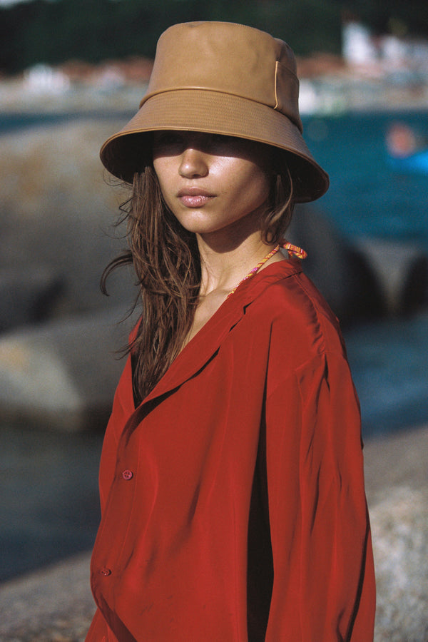 Model wearing a tan vegan leather bucket hat with a red shirt by the beach
