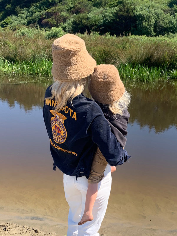 A child wearing a brown Teddy Bucket hat, held by an adult in a natural setting by a river.