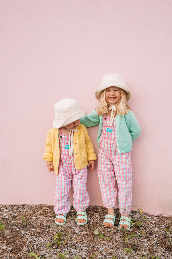Two children wearing colorful outfits and beige bucket hats against a pink wall.