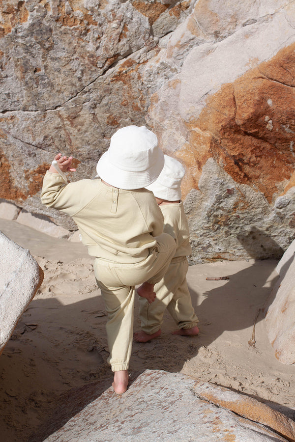 Two children wearing beige cotton bucket hats playing on a beach near rocks.