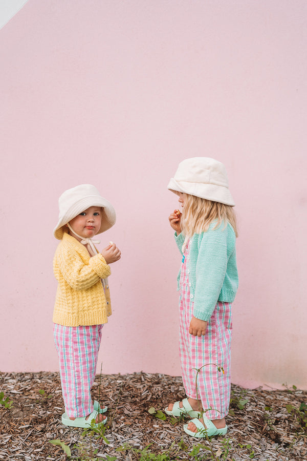 Two children wearing beige bucket hats, one in yellow and one in green, against a pink wall.