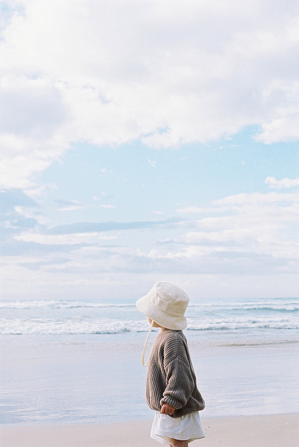 Child wearing a beige cotton bucket hat on the beach, looking at the ocean under a cloudy sky.