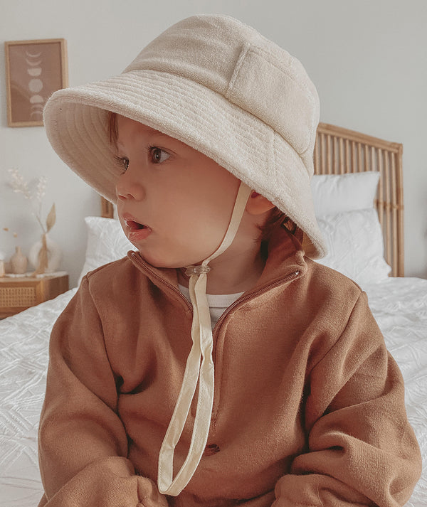 Child wearing a beige cotton bucket hat with neck tie, sitting on a bed