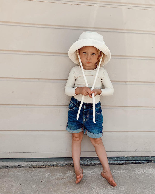 Child wearing a beige cotton bucket hat with straps, standing on a concrete surface.